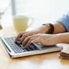 hands typing on a laptop computer on a wooden desk next to a cup of tea