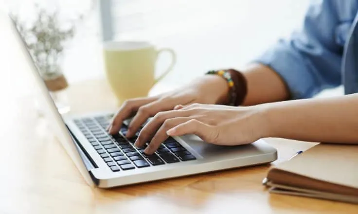 hands typing on a laptop computer on a wooden desk next to a cup of tea