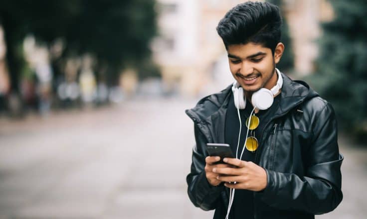 male blogger in leather jacket typing on his cell phone while walking on an empty street