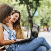 2 women sitting on a bench in the park smiling at a laptop