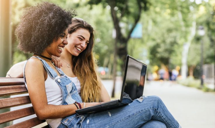 2 women sitting on a bench in the park smiling at a laptop