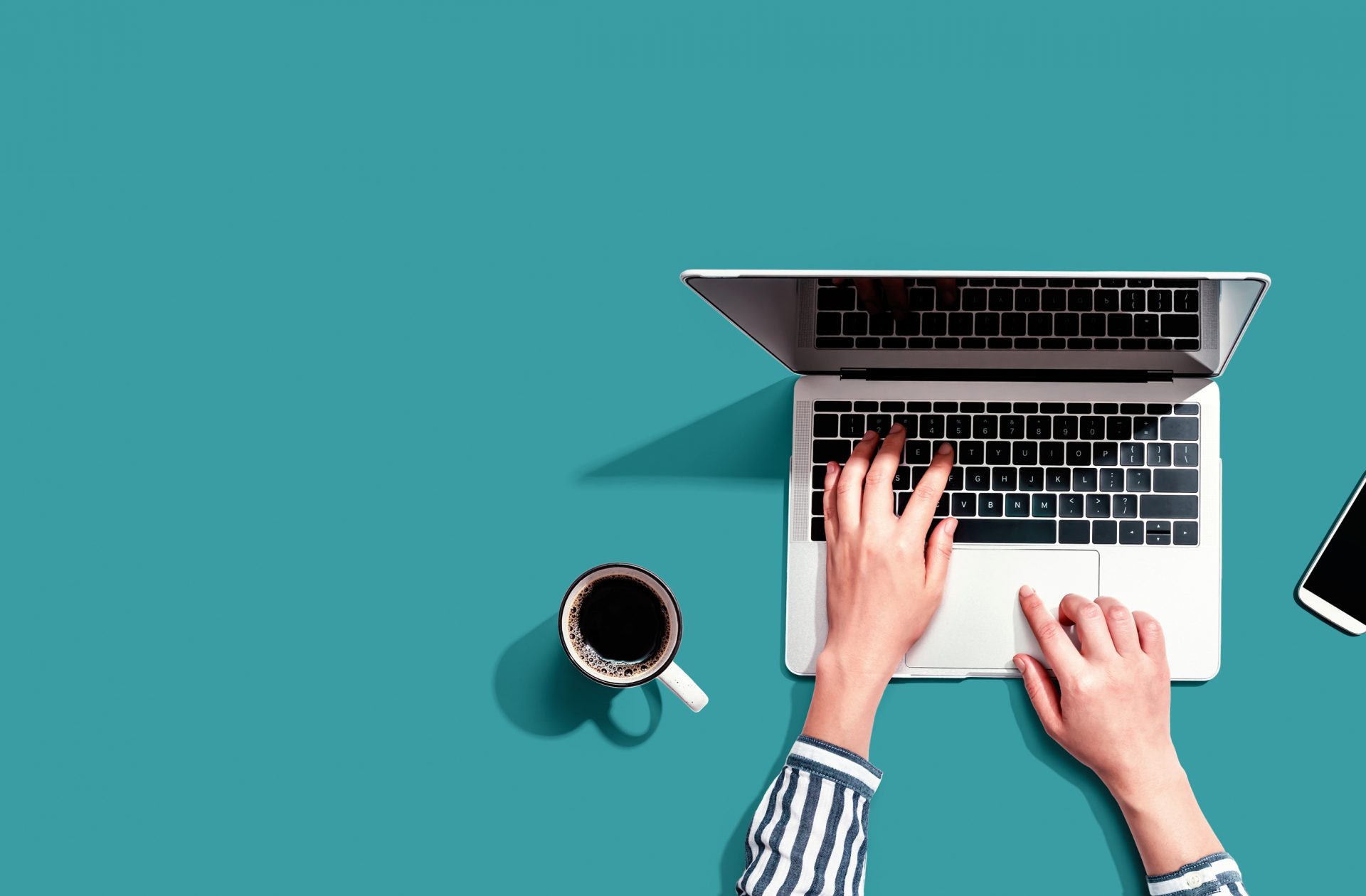 hands of a woman in a striped blouse typing on a silver laptop with a smartphone and cup of coffee next to her on a teal background