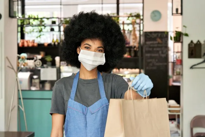 female restaurant worker wearing a mask and handing a brown paper bag to a customer