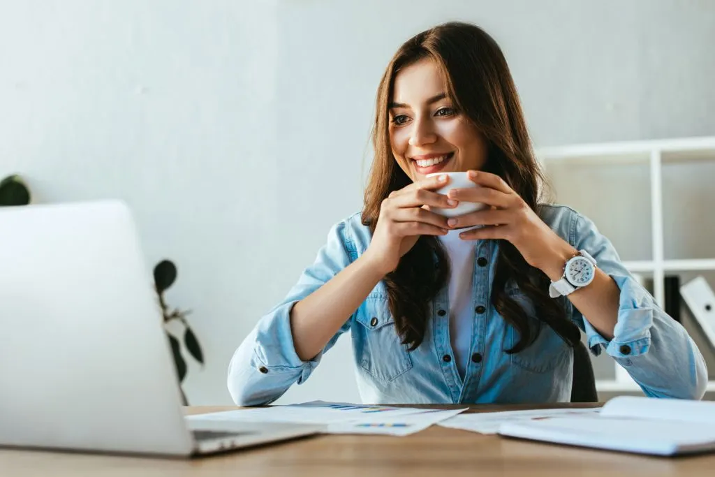 woman smiling at a laptop and holding a cup of coffee