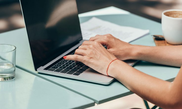 woman's hands typing on a laptop on a blue patio table with a cup of coffee and a stack of papers nearby