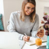 a blogger writes in her planner while looking at her phone and computer on a white desk with a teal vase of purple flowers.