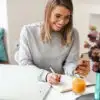 a blogger writes in her planner while looking at her phone and computer on a white desk with a teal vase of purple flowers.