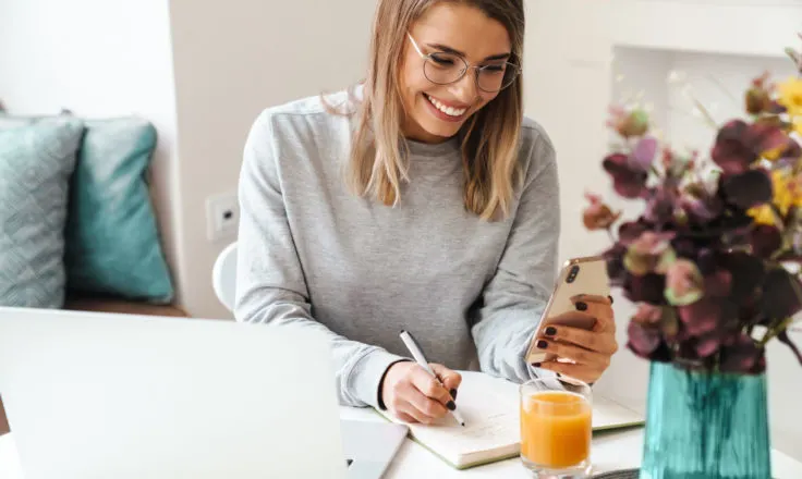 a blogger writes in her planner while looking at her phone and computer on a white desk with a teal vase of purple flowers.