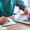 person writing on notebook and using a laptop