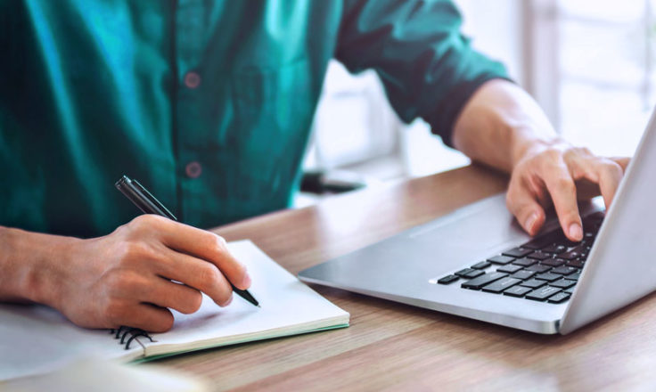 person writing on notebook and using a laptop