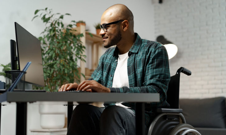 man in a wheelchair wearing a plaid shirt working on a computer at a desk