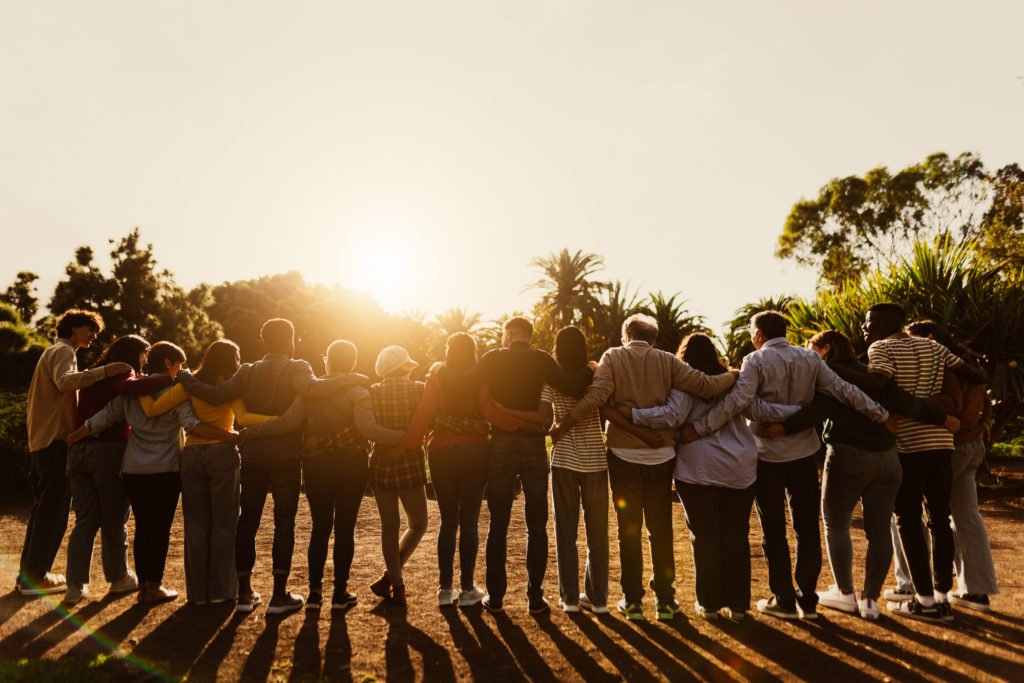 group of multigenerational people standing in a park with linked arms at sunset with their backs facing the camera