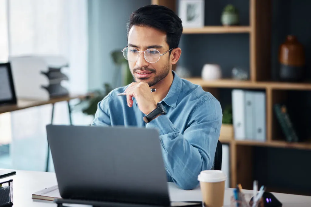 image of a man wearing glasses and a blue shirt sitting at a desk working on a laptop